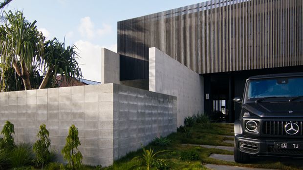 Street-facing view of a contemporary residence with a dark vertical-screen facade, exposed concrete block boundary walls, a recessed entry or carport, and tropical landscaping. A vehicle is parked in the foreground, while the building's strong geometric forms and layered screening dominate the composition.