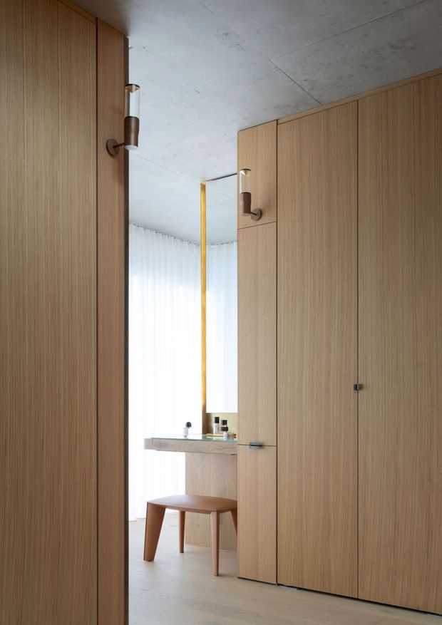 A refined built-in wardrobe or dressing area with full-height, vertically grained light timber cabinetry and discreet dark hardware. A narrow opening reveals a bright vanity zone with a stone benchtop, integrated mirror, stool, and sheer curtains. The ceiling is raw concrete, creating a warm minimalist interior with subtle industrial architectural detailing.