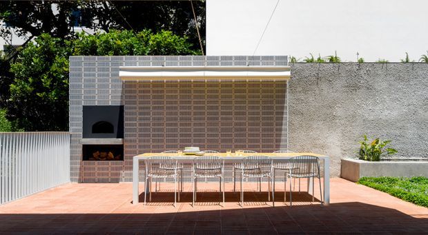 A sunny contemporary outdoor dining and cooking area with a long dining table and woven chairs on a terracotta-toned tiled terrace. Behind it is a built-in barbecue or fireplace set into a patterned breeze-block screen, with a white rendered wall, raised planter, and lush greenery. The space has strong indoor-outdoor entertaining character and crisp modern detailing.