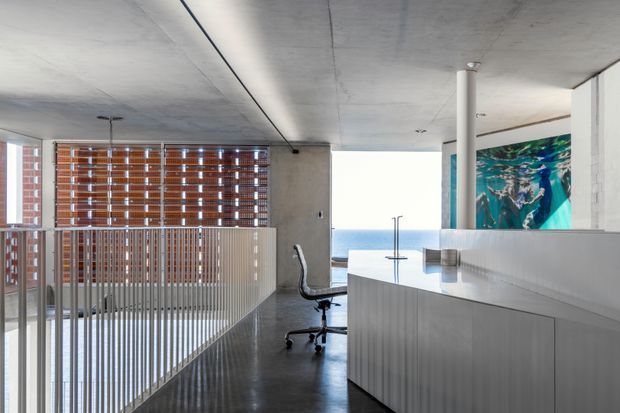A contemporary open-plan interior with polished dark flooring, exposed concrete ceiling, white built-in cabinetry, and a workspace overlooking the ocean. Vertical metal balustrades screen a stair or void area, while timber venetian blinds filter light through large windows. A colorful aquatic artwork is mounted above the cabinetry, and a bright ocean horizon is visible beyond the glazing.