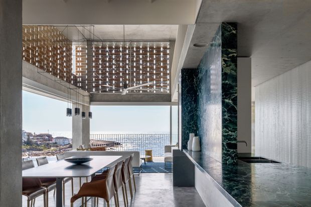 A sophisticated open-plan dining and kitchen interior in a concrete coastal residence. A long dining table with upholstered chairs sits beside full-height glazing overlooking the ocean, while a dark green marble-clad kitchen island and matching vertical feature wall dominate the right side. Exposed concrete ceilings and columns, timber screening, linear pendant lights, and restrained contemporary furnishings create a refined, airy setting.