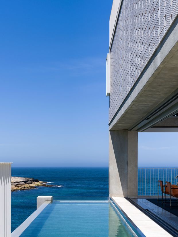 A contemporary coastal building exterior overlooking the ocean, with a rectangular infinity-edge pool in the foreground and a covered terrace supported by substantial concrete columns. The upper facade is clad in a pale patterned masonry or brick screen, while the terrace has metal-framed glazing and a slender metal balustrade. Bright blue sky and deep blue sea create a strong coastal setting.