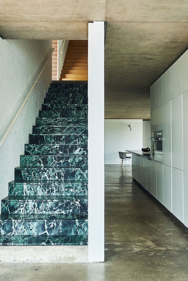 Interior view of a contemporary open-plan kitchen and stairway. A dramatic dark green veined stone staircase dominates the foreground, rising beside a white-painted brick wall and raw concrete ceiling. The kitchen beyond has sleek full-height pale grey cabinetry, integrated appliances, a dark island bench, and a polished or sealed concrete floor. The composition highlights the contrast between the sculptural stone stair and restrained industrial-style kitchen fit-out.