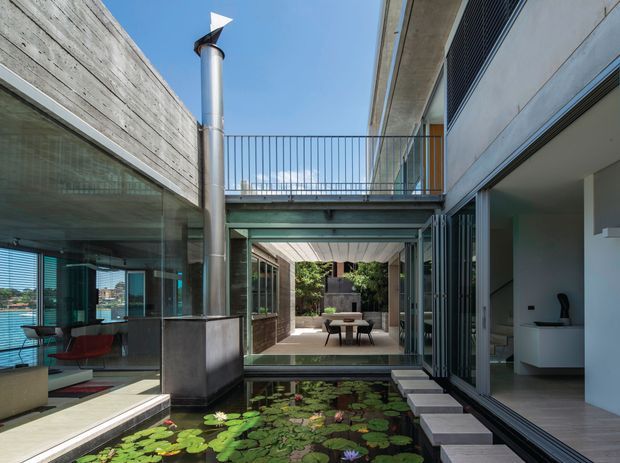 A contemporary multi-level courtyard viewed from within a modern residence, centred on a shallow lily pond with stepping stones. The space features exposed concrete, extensive glazing and sliding glass doors, a steel-framed upper bridge and balcony railing, timber-lined covered areas, and a visible outdoor dining setting. An interior living or dining area opens directly onto the courtyard, while a sculptural metal flue rises beside the glazing.