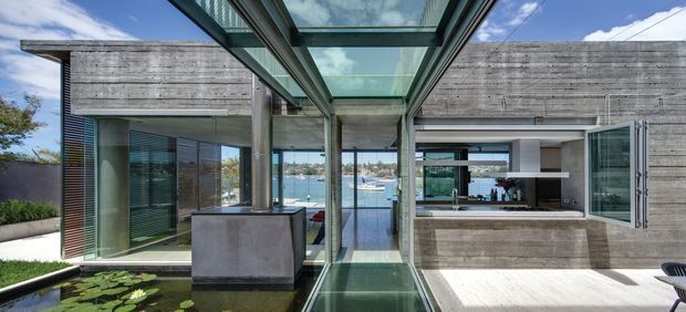 Contemporary waterfront courtyard view of a concrete-clad residence, centred on a glazed overhead and walkway structure. The image shows exposed concrete walls, extensive floor-to-ceiling glazing, a modern outdoor kitchen or servery with cabinetry, and a reflective water feature with lily pads in the foreground. Boats and water are visible beyond the house through the glazing.
