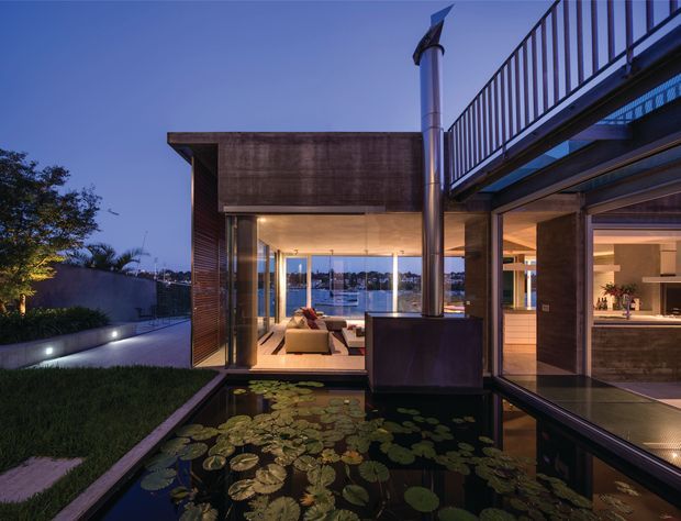 Twilight view of a refined modern residence arranged around a reflective lily pond and landscaped courtyard. The composition features warm timber-clad walls, extensive floor-to-ceiling glazing, a covered living area visible inside, an open-plan kitchen or bar area to the right, and a metal roof terrace or balcony with railing above. A sculptural vertical chimney or flue rises through the courtyard, while distant water is visible beyond the glazing.
