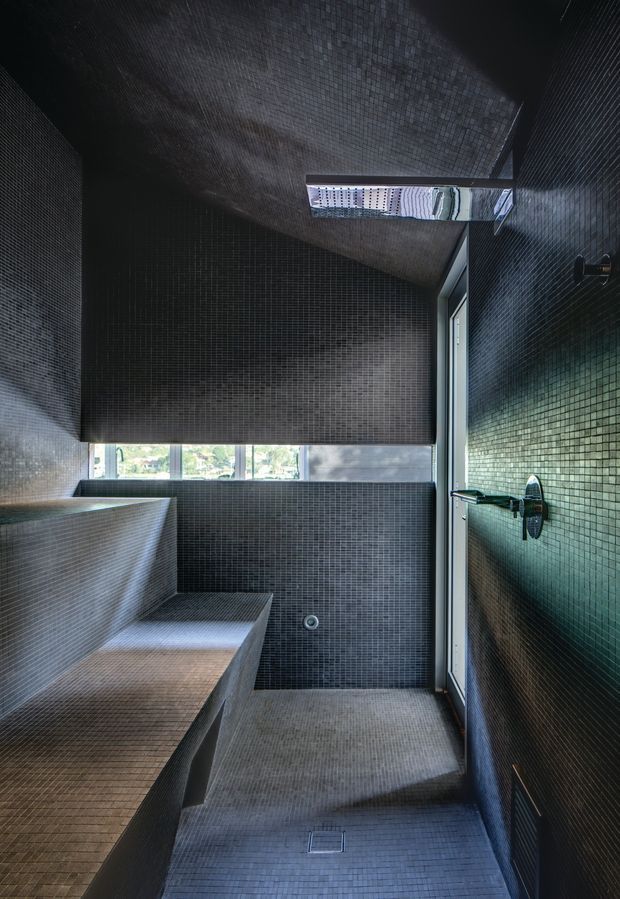 A dark, compact tiled bathroom or wet-room with charcoal mosaic tiles covering the walls, floor, ceiling, and built-in bench-like surfaces. A wall-mounted shower fixture is visible on the right, with a narrow skylight above and a small horizontal window bringing in daylight. The space has a highly monolithic, minimalist architectural character and a moody grey-black palette.