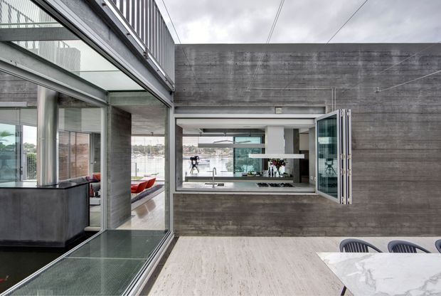 Contemporary open-plan interior with exposed concrete walls and columns, extensive floor-to-ceiling glazing, and a large open window connecting the dining area to an adjacent kitchen or living space. A marble dining table with modern chairs occupies the foreground, while a kitchen island and colourful seating are visible through the glazing and opening. The space has a refined industrial character, abundant natural light, and strong indoor-outdoor visual connections.
