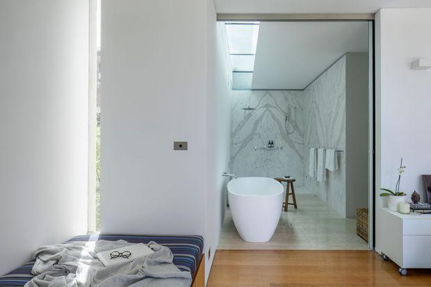 A bright contemporary bedroom opens directly into a refined bathroom. The bedroom has white walls, warm timber flooring, and a low striped upholstered bed or daybed in the foreground. The bathroom features large pale marble-look wall and floor surfaces, a freestanding white oval bathtub, wall-mounted shower fittings, skylight glazing, and minimal white cabinetry. The space is airy, clean, and softly luxurious.