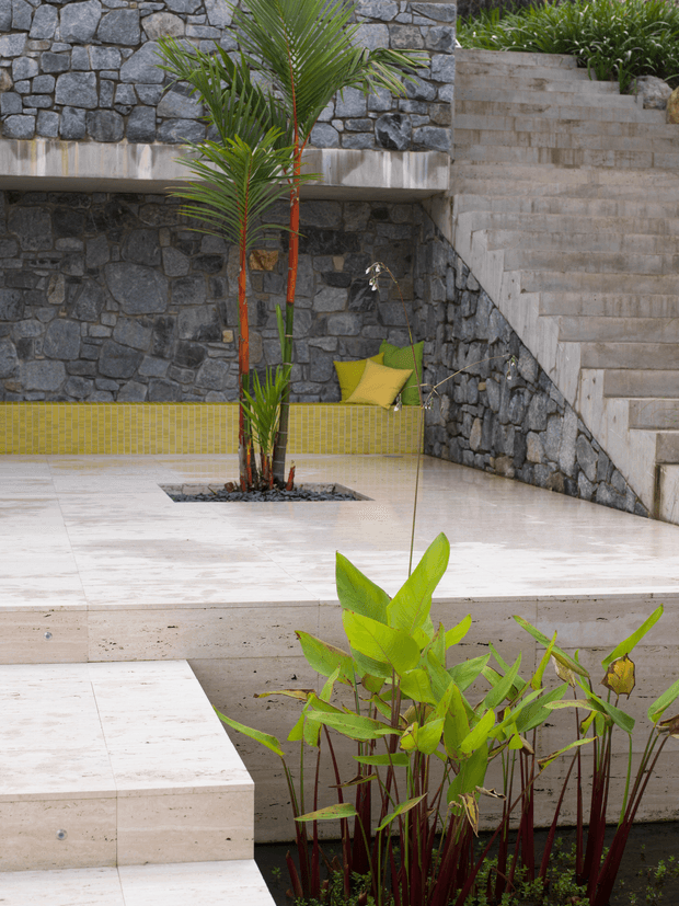 A landscaped outdoor courtyard or pool terrace featuring dark irregular stone masonry walls, pale stone paving and steps, a yellow-green tiled built-in bench, tropical palms and planting, and a small hanging chair. The design has a layered, resort-like feel with strong geometric stair and platform forms.