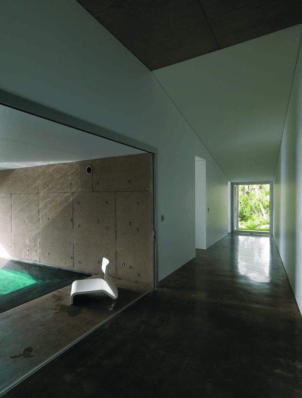 A moody modern interior corridor with polished dark concrete flooring, smooth pale walls and a dark concrete ceiling plane. The hallway opens to a partially visible indoor pool area behind a large glazed opening, with a sculptural white chair beside the pool. Bright greenery is visible through the end glazing, creating a strong contrast with the dark, minimal interior.