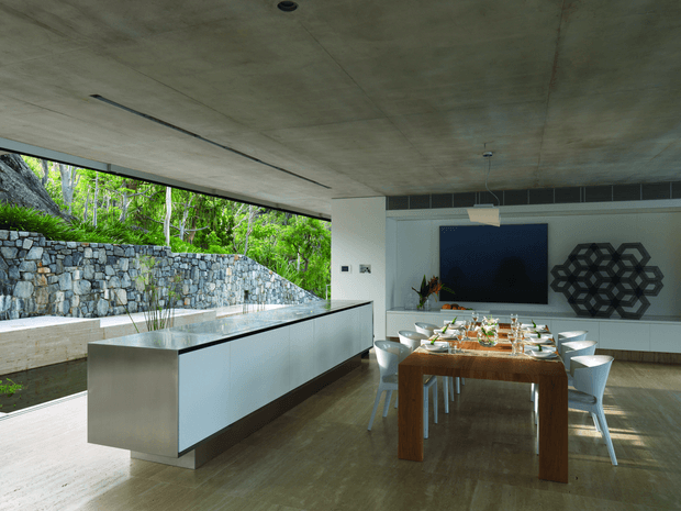 A contemporary open-plan dining and entertaining space with a long timber dining table set for a meal, surrounded by white modern chairs. A large white island or built-in counter with a dark stone-like top occupies the foreground. The room features a raw concrete ceiling, pale walls, timber flooring, concealed linear air-conditioning, a large dark blue wall panel, and geometric dark wall decor. Full-height open glazing connects the interior to a landscaped stone retaining wall and lush greenery outside.