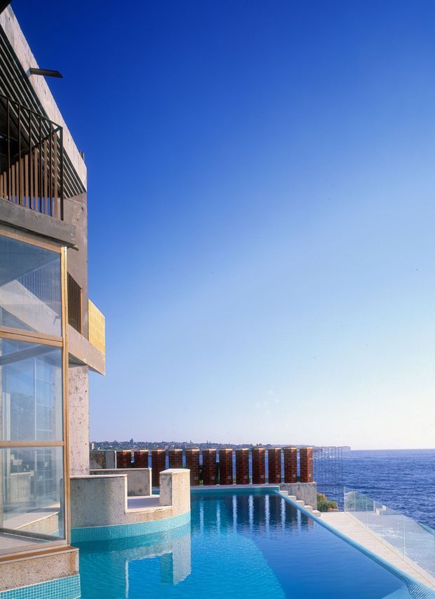 A modern multi-level coastal building with exposed concrete walls, extensive floor-to-ceiling glazing, timber-toned vertical screens and balconies. The image focuses on a turquoise swimming pool terrace overlooking the ocean, with a clear blue sky and distant coastline visible beyond.