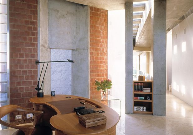 A refined open-plan interior featuring a curved timber desk or built-in worktable in the foreground, exposed red-brick and raw concrete walls and columns, polished pale stone flooring, and a narrow integrated bookshelf. Strong natural light enters through vertical glazing and a linear overhead opening, creating a gallery-like workspace with a clear view toward an adjoining circulation area.