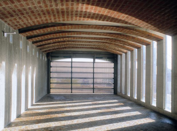 A long, enclosed architectural passage or garage-like space with a curved exposed brick-vaulted ceiling, repeated dark metal structural beams, rough concrete side walls, tall narrow openings along one side, and a metal gate at the far end. Strong sunlight creates repeated linear shadows across the textured floor, giving the space an industrial, historic, and dramatic character.