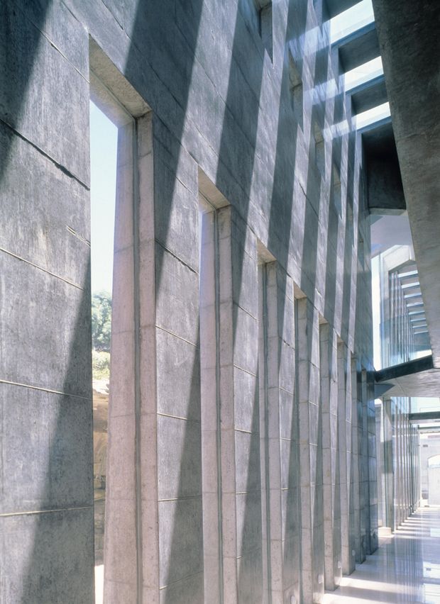 A monumental architectural passage or colonnade lined with tall rectangular concrete piers and large recessed openings. Strong daylight enters through the openings and overhead glazing, creating dramatic linear shadows across the textured grey concrete surfaces. A glazed walkway and additional structural bays recede into the distance, emphasizing the building's vertical rhythm and geometric form.