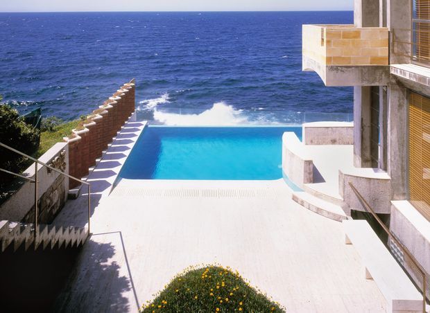 Exterior view of a modern multi-level coastal residence with a vivid rectangular swimming pool overlooking the ocean. The scene features pale stone or concrete terraces, projecting balconies, horizontal timber or metal sun-screen blinds, a textured brick privacy wall, and a broad pool deck descending toward the water. Bright sunlight and expansive ocean views create a dramatic seaside setting.