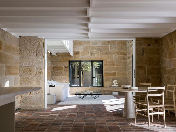 A warm, light-filled open-plan interior with exposed sandstone block walls, a dark terracotta-toned tiled floor, white exposed ceiling beams, and a dining area beside a compact kitchen. Black-framed glazing opens toward a bright courtyard-like space, while a white sculptural dining table and timber chairs provide a contemporary focal point.