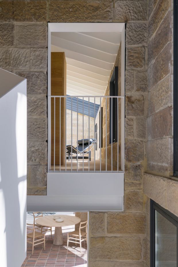 A vertical architectural interior view through a narrow opening framed by rough-cut sandstone masonry. A white-painted projecting balcony or mezzanine with slim metal balustrade overlooks a lower dining area. Beyond it is a bright corridor or courtyard-like passage with timber joinery, exposed stone walls, extensive glazing, and a white slatted ceiling. The image highlights layered spatial connections, natural light, stone construction, and a contemporary renovation aesthetic.