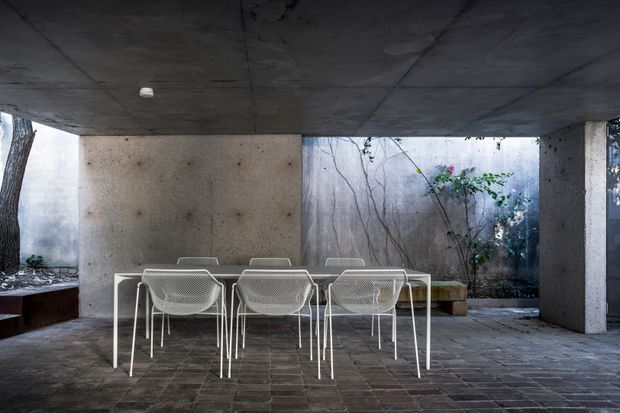 A moody, minimalist covered dining terrace or courtyard with a long white dining table and matching mesh chairs. Raw concrete ceiling, walls, columns and openings frame a planted courtyard with exposed tree branches, while dark rectangular masonry or stone paving covers the floor. The space has a sheltered, indoor-outdoor character and strong geometric composition.