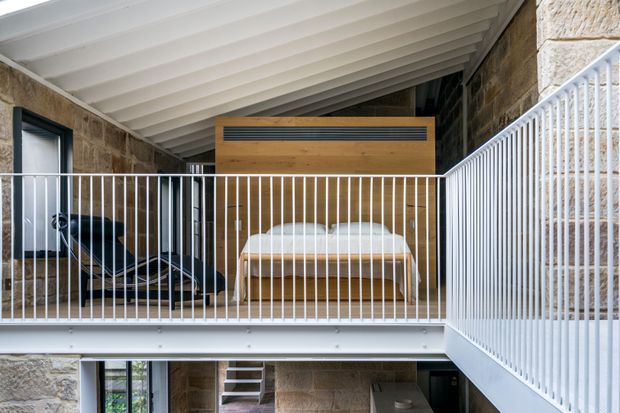 A sheltered elevated terrace or balcony within a stone-clad building, featuring a white metal balustrade, a timber-clad built-in element, outdoor lounge seating, and a white slatted ceiling. Stone walls and glimpses of stairs and interior spaces create a refined contemporary architectural setting.