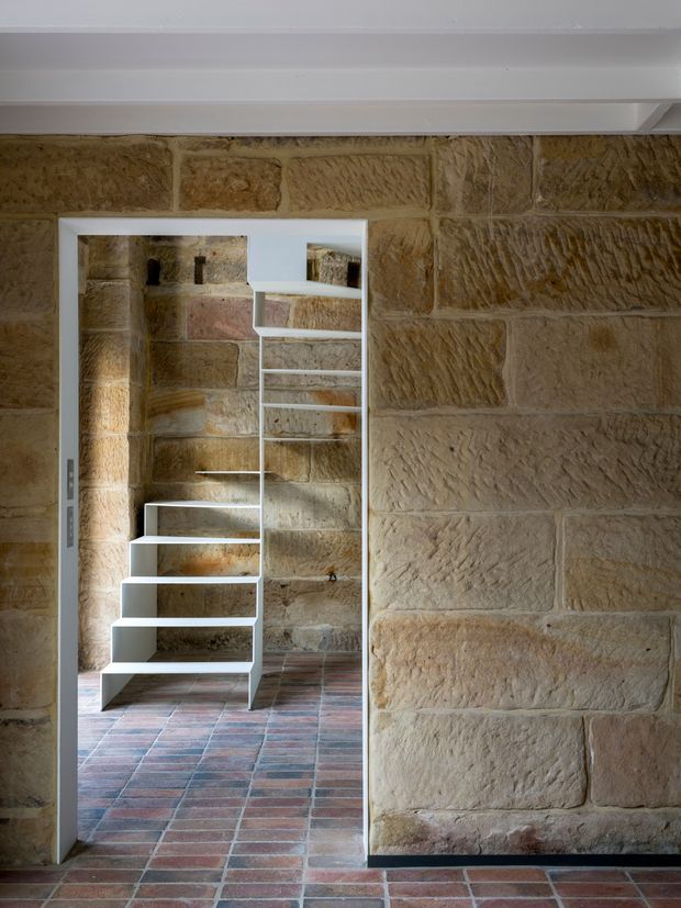 Interior architectural detail showing a narrow doorway opening in a heavily textured sandstone block wall, leading to a compact modern open-riser stair. The stair has crisp white stringers and treads, while the adjoining room continues the exposed stonework and includes a reddish-brown tiled floor. A white ceiling beam or bulkhead spans the top of the image. The composition contrasts historic masonry with contemporary stair construction.