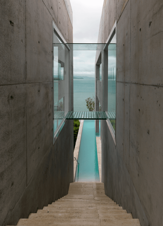 A dramatic architectural view down a long concrete stairway between tall raw concrete walls. A narrow turquoise swimming pool extends beyond the bottom of the stairs toward a glass-enclosed bridge and expansive ocean view. The composition features exposed concrete, extensive glazing, a strong axial perspective, and a restrained modern palette.
