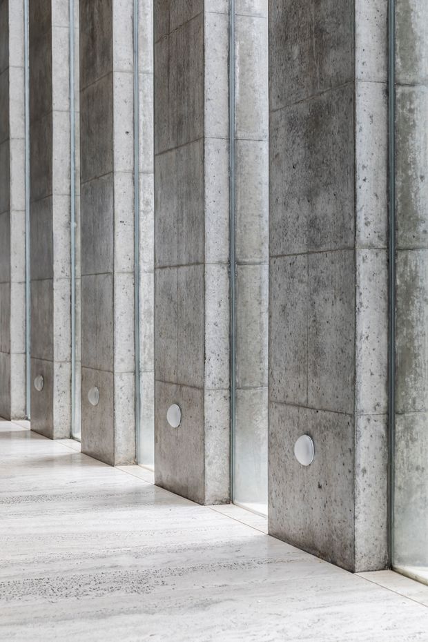 Close-up architectural view of a repetitive interior wall made from raw, board-formed concrete piers separated by tall narrow glazed panels. Small round wall lights are embedded near floor level, and the pale polished concrete floor continues along the corridor-like space. The composition is monochromatic, textured, and strongly geometric.
