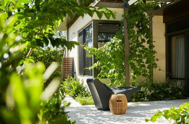 A leafy, private outdoor courtyard or terrace beside a contemporary home. Pale stone or concrete paving forms the main surface, with dense green planting and climbing vines around large dark-framed windows and a covered structure. Two sculptural dark outdoor lounge chairs and a small rounded slatted timber side table create a relaxed seating area. The image is sunlit, lush, and tranquil.