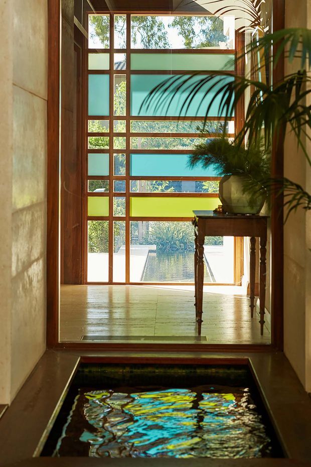 A narrow, warmly lit interior passage leads toward a tall timber-framed glazed screen and exterior garden water feature. The glazing incorporates horizontal and vertical coloured translucent panels in turquoise, green, and pale blue, with views of foliage and water beyond. A traditional dark wood console table with a large potted plant stands to the right, while a shallow reflective indoor water feature occupies the foreground. The space has polished stone or tile flooring, warm timber framing, and earthy plastered walls, creating a serene, nature-connected atmosphere.