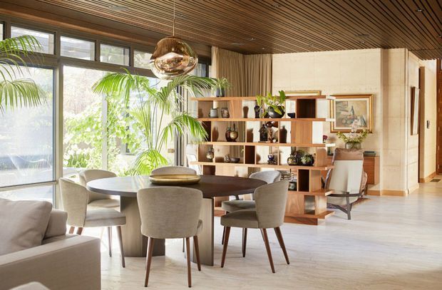 A bright, refined open-plan dining and living area with a large round dining table surrounded by upholstered chairs. Floor-to-ceiling glazing brings in abundant daylight and greenery, while a sculptural pendant hangs above the table. A timber open display shelving unit separates the dining zone from a compact sitting or study area, and the room features a warm slatted timber ceiling, pale flooring, and neutral furnishings.