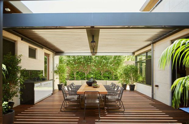 A contemporary covered outdoor dining terrace framed by a dark steel and timber structure. A long wooden dining table with multiple wire-frame chairs sits on a timber deck beneath a slatted pergola, with integrated lighting, pale rendered walls, extensive glazing, and lush planted greenery beyond. The space is bright, open, and indoor-outdoor in character.