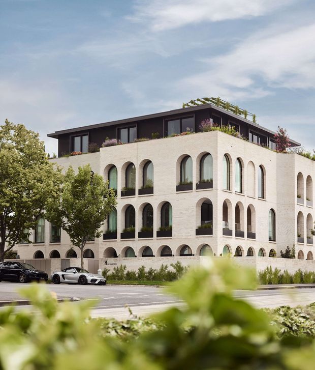 Exterior view of a contemporary multi-storey building with a pale brick facade, repeated tall arched windows and openings, dark upper-level cladding, and planted rooftop terraces. The building is set along an urban street with landscaped planting in the foreground and cars at the curb.