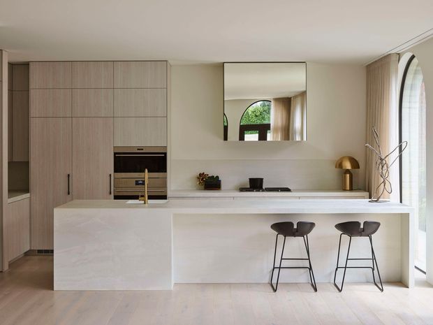 A bright, refined contemporary kitchen with a large pale stone island and integrated breakfast bar. The kitchen features light timber floor-to-ceiling cabinetry, concealed appliances, a slim brass-toned tap, minimalist white walls and benchtops, and two dark sculptural bar stools. A large mirror reflects an arched glazed opening, while a tall arched window with sheer curtains brings in abundant natural light. Decorative objects and a brass dome lamp add restrained detail.