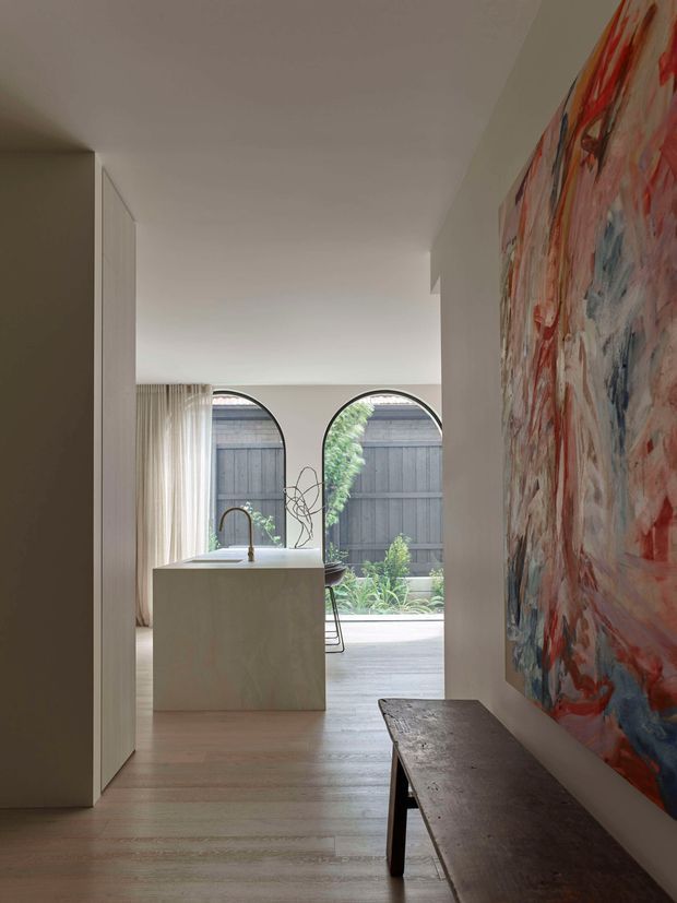 A refined interior view looking through a minimalist hallway toward an open-plan kitchen. Pale timber flooring, white walls and streamlined built-in cabinetry create a calm, contemporary setting. The kitchen features a large light-coloured island with a curved brass-toned tap, while arched windows frame a planted courtyard beyond. A long dark timber bench and a large expressive abstract artwork add contrast and visual interest along the hallway wall.