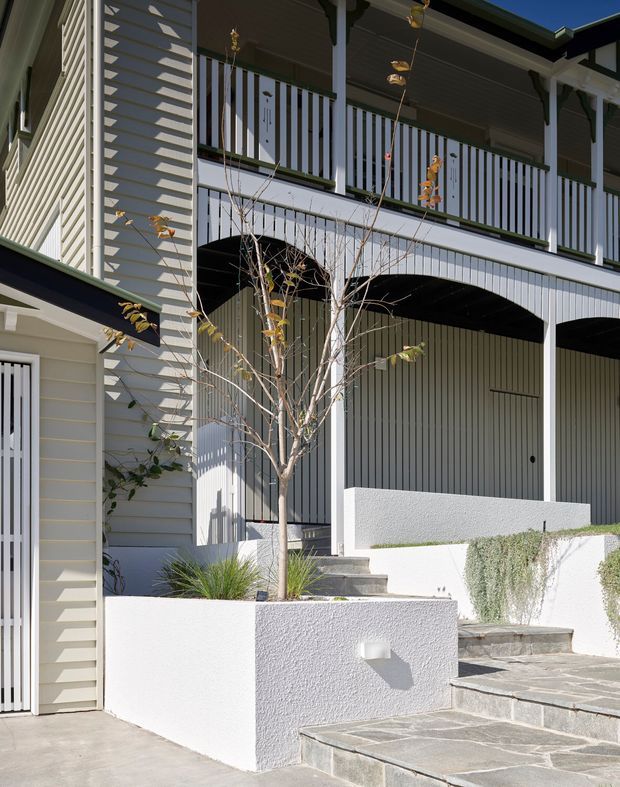 Exterior view of a multi-level residential building with pale horizontal weatherboard cladding, a covered carport or entry area, and an upper balcony with white vertical balustrades. White rendered planter walls, grey stone-paved steps, and restrained landscaping lead toward the entrance. Strong sunlight creates crisp shadows across the facade and balcony.