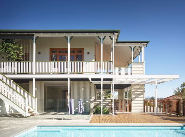 Bright exterior view of a two-storey residential house with a broad covered upper veranda, white balustrades, timber-framed doors and windows, vertical light cladding, external stairs, a lower timber deck and a swimming pool in the foreground. The structure has a layered pitched roof and open outdoor entertaining areas.
