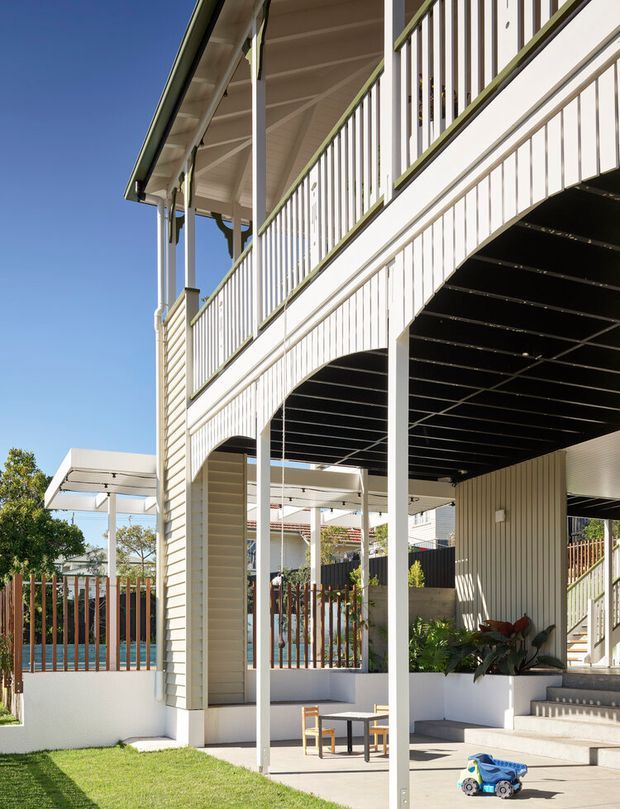 Bright exterior view of a raised, two-level residential house with a covered ground-floor patio and upper verandah. The facade uses light painted horizontal weatherboards, white vertical balustrades and posts, and a pitched roof with exposed underside framing. Outdoor stairs lead to the upper level, while the lower terrace includes a small table, chairs, planting, and a fenced swimming pool area visible beyond.