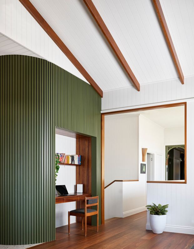 Bright interior featuring a compact built-in study nook with timber desk, shelving, books and chair set into a dramatic curved wall clad in dark green vertical battens. White vertical wall panelling and a white vaulted ceiling with exposed warm timber beams create a light, refined character. A stair opening and large timber-framed interior opening are visible to the right, with timber flooring throughout and a potted plant near the stair.