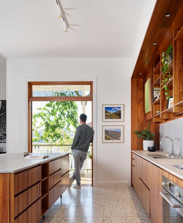 Bright galley-style kitchen with a central island, warm timber cabinetry and open shelving, white vertical wall panelling, a white paneled ceiling, and a terrazzo floor. The kitchen opens to a balcony or exterior view through a glazed timber-framed door, with integrated appliances, a sink, framed artwork, and indoor plants.