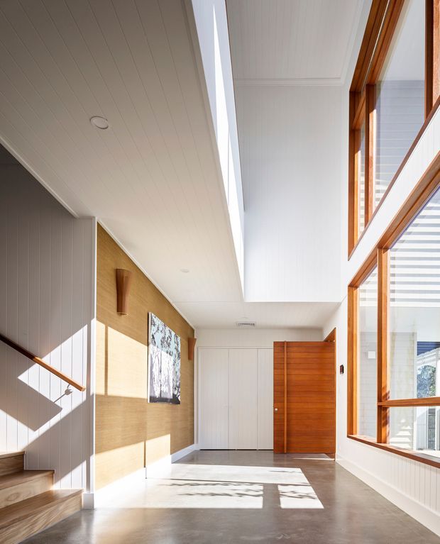 Bright, spacious residential entry hall with a polished concrete floor, white vertically panelled walls and ceiling, a warm timber feature wall, and a timber pivot front door. A stair rises at the left with a simple timber handrail, while large timber-framed windows on the right bring in strong natural light and overlook an exterior structure. The hall has a high ceiling with a long clerestory-like opening, clean-lined built-in doors, and a restrained contemporary coastal feel.