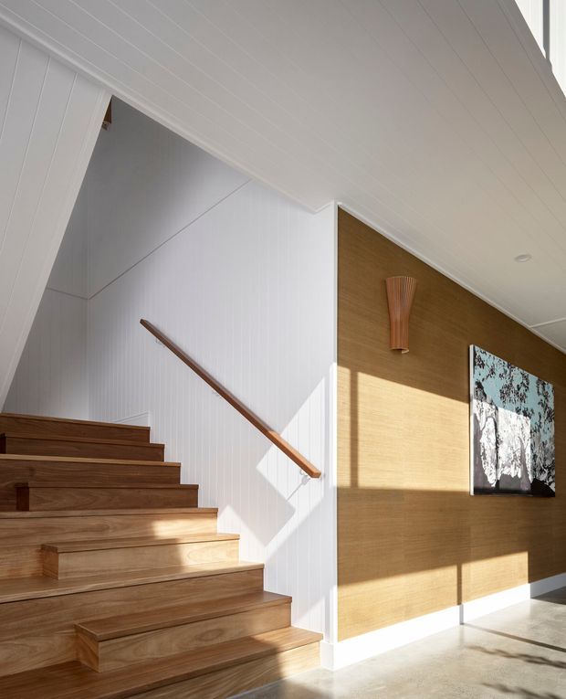 Bright contemporary stair hall with broad timber stairs, a white vertically panelled stair enclosure, and a warm timber feature wall. A slim timber handrail follows the staircase, while a sculptural wall light and framed artwork add decorative detail. Strong sunlight casts angular shadows across the walls and pale floor.