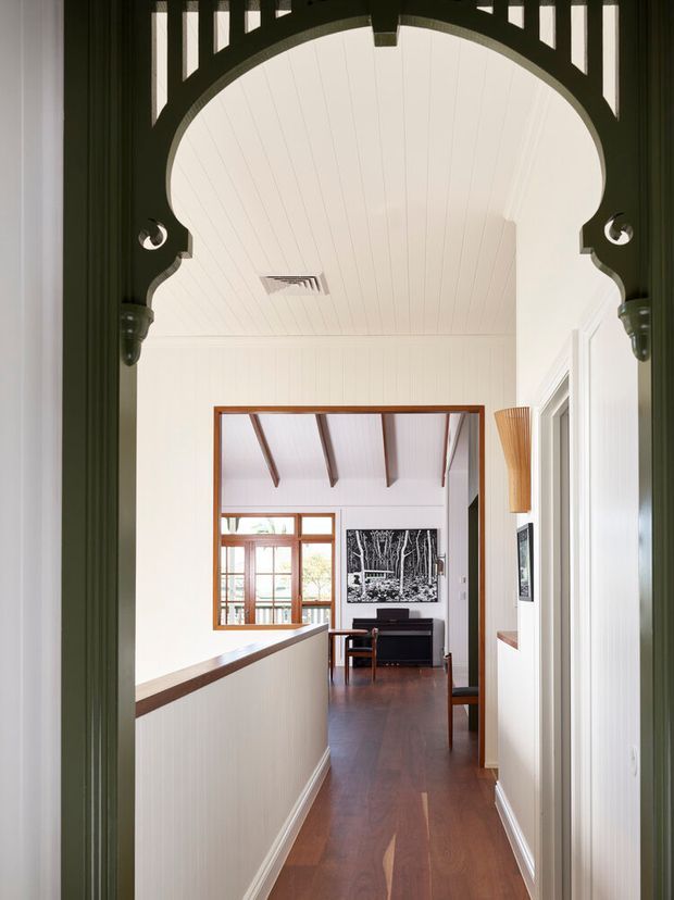 A bright, narrow residential hallway viewed through an ornate dark-painted archway. The corridor has white vertical wall panelling, a white timber-lined ceiling, and polished dark timber flooring. At the far end, the hallway opens toward a small sitting or living area with exposed timber rafters, timber-framed glazing, a dark console or piano-like cabinet, and framed artwork. The image combines period decorative detailing with a clean, light interior and warm timber accents.