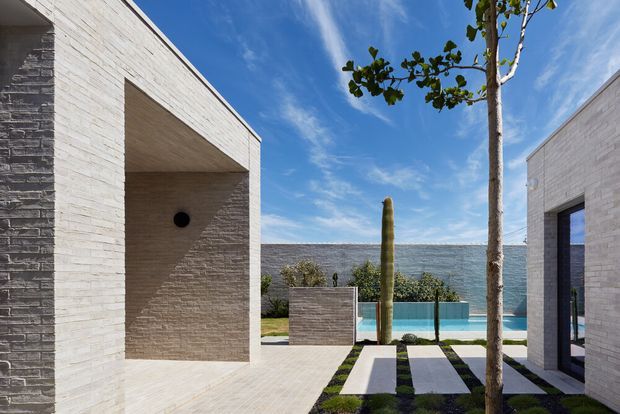 Exterior architectural view of a contemporary, rectilinear property arranged around a landscaped courtyard and pool. Pale textured masonry walls form deep recessed openings and framed views toward the water, with a paved path, clipped planting, slender trees, and a vivid blue sky. The composition emphasises strong geometric forms, light stone-like brickwork, and indoor-outdoor connection.