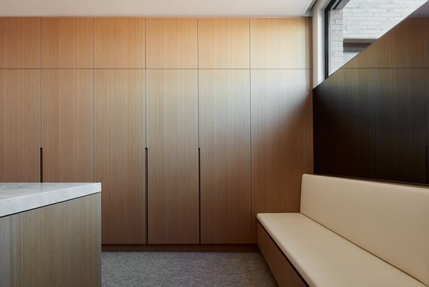 A refined contemporary interior featuring full-height warm timber wall cabinetry, a pale stone island or counter at left, and a built-in cream upholstered bench seat at right. Dark timber panelling and a high window add contrast, while grey carpet and clean linear detailing create a calm, minimalist look.