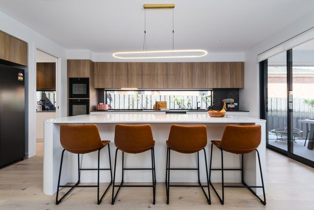 Contemporary open-plan kitchen dominated by a large white island with four brown upholstered bar stools. Warm timber cabinetry spans the rear wall, with integrated black appliances, a dark benchtop and splashback area, and a full-width window above the work zone. White walls and ceiling, pale timber flooring, black-framed glazing, and a linear oval pendant light create a bright, refined interior. A glazed outdoor area is visible to the right.