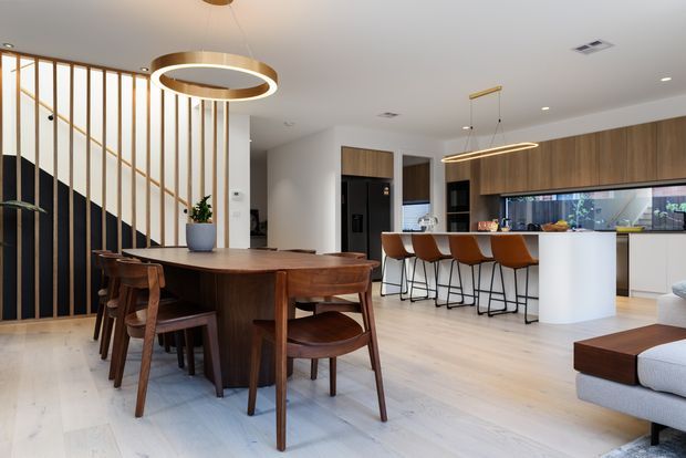 Open-plan contemporary kitchen and dining area with a dark timber dining table and chairs, pale timber flooring, a white island with seating, warm timber cabinetry, and a black vertical-slat feature screen separating the dining space from the stairway. Linear and circular pendant lighting provide warm accents.