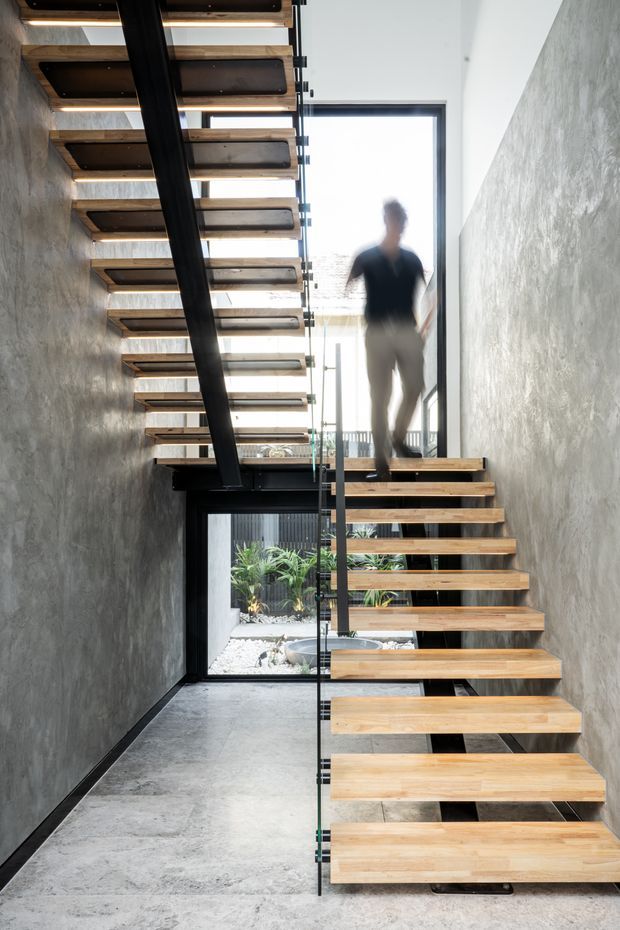 A contemporary interior stairway with an open-riser floating staircase. Thick timber treads are supported by a dark metal central structure and paired with a frameless glass balustrade. The stairwell has textured grey concrete-like walls, a polished concrete floor, large glazed openings, and a small planted courtyard visible beyond. A blurred person is walking on the stairs.