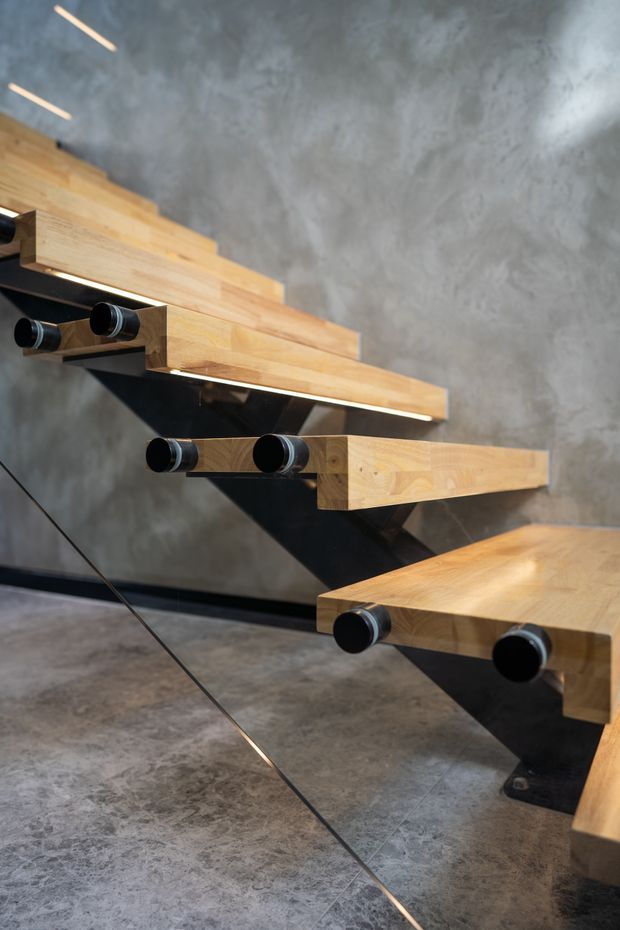 Close-up architectural photograph of a sculptural floating staircase with thick light timber treads, black steel support brackets, and cylindrical black metal detailing. The staircase is set against a textured grey concrete wall and floor, with integrated linear lighting beneath the treads and a glass balustrade edge visible in the foreground. The composition emphasizes the dramatic cantilevered construction and material contrast.