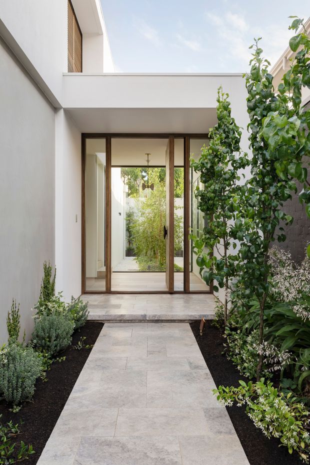 A bright, minimalist courtyard entry viewed from an exterior garden path. Large pale stone pavers lead through landscaped planting to full-height glazed timber-framed doors. White rendered walls, a flat-roofed overhead structure, and a compact open-air courtyard create a clean indoor-outdoor connection, with a glimpse of greenery and a further garden beyond the doors.